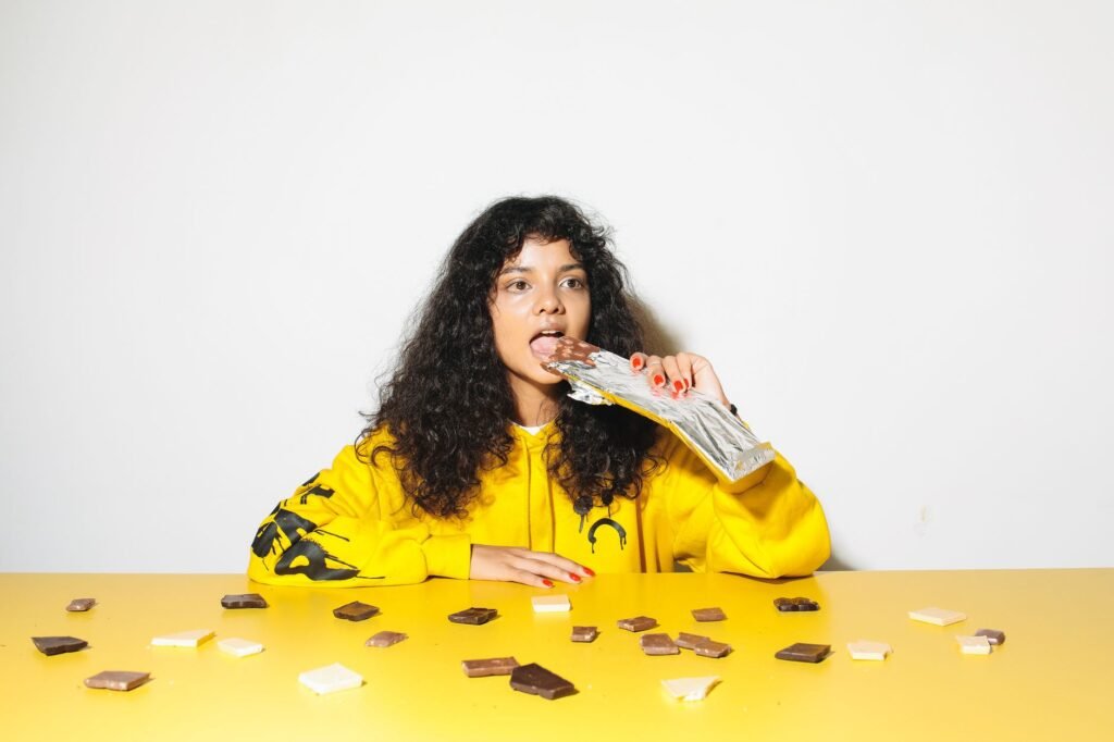 A young woman in a yellow hoodie enjoying chocolate pieces at a table. Bright scene.
