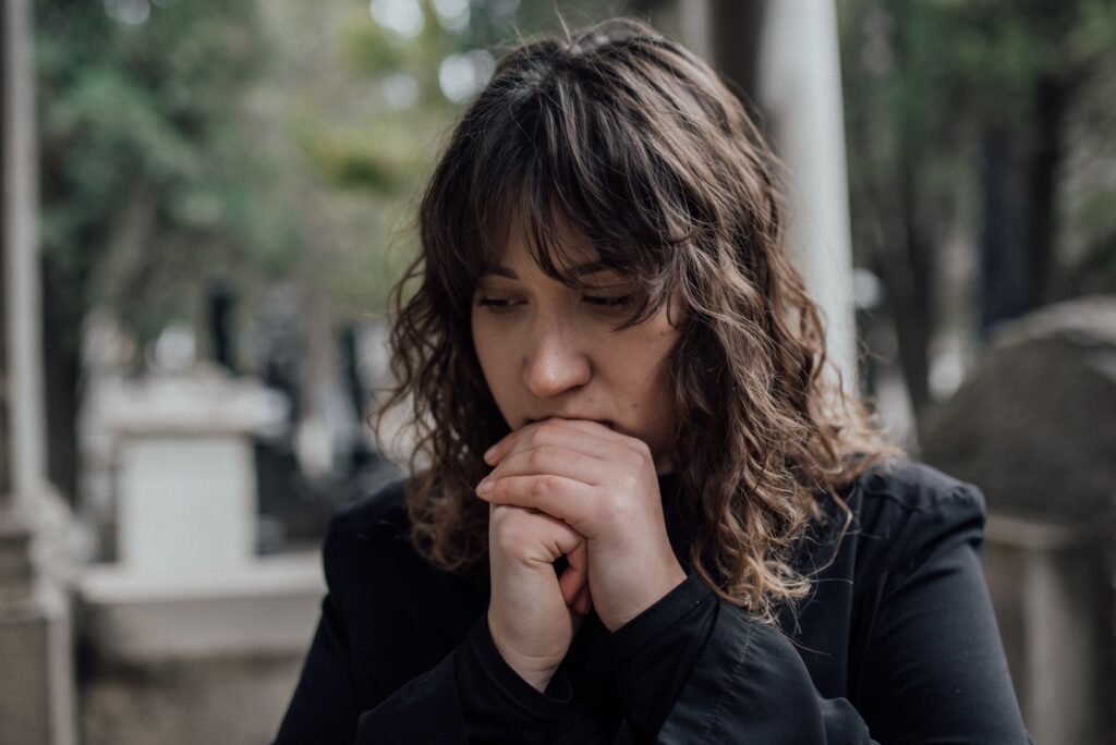 Pensive woman in black coat standing outdoors with hands together, conveying sadness.