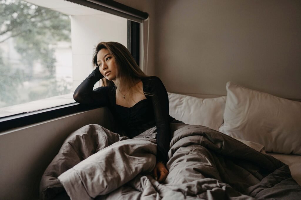 A young woman in a cozy room, pondering by the window, wrapped in blankets.